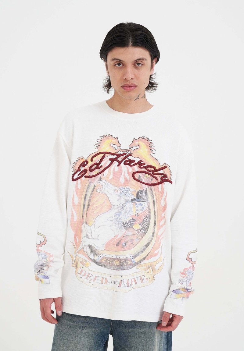 Jonge man met schouderlang donker haar draagt een witte lange mouwen shirt met vlammenpaarden, een skeletrijder en de teksten "Ed Hardy" en "Dead or Alive".