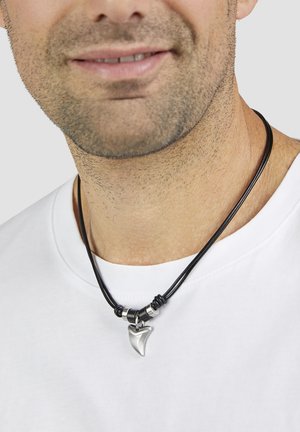 Schwarze Lederkette mit einem silbernen, zahnförmigen Anhänger und kreisförmigen metallischen Akzenten. Getragen über einem weißen Hemd.