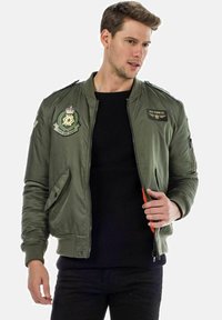 Olijfgroene bomberjack met geborduurde patches, geribbelde kraag, zijzakken en oranje ritsdetails, gedragen over een zwarte gebreide trui.