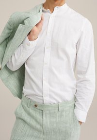 Man i vit skjorta med knappar som håller en ljusgrön strukturerad kostymjacka över axeln, klädd i matchande ljusgröna strukturerade byxor.