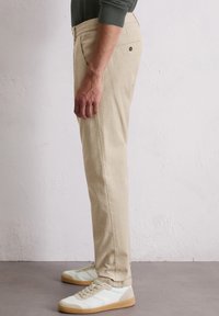 Un homme portant un pantalon beige et des baskets blanches se tient de côté contre un mur blanc uni et un sol gris.