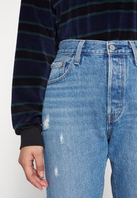 Vaqueros de tiro alto azules con detalles desgastados, que presentan un diseño clásico de cinco bolsillos y cierre de botón de metal. El material parece denim.