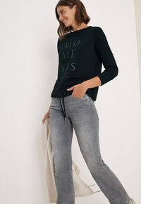 Donkergroen pullover met grafische tekst "MOMENTS" in een lichter groen, gecombineerd met lichtgrijze jeans en een off-white cardigan.