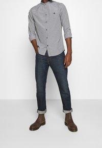 Camisa de botones con patrón de cuadros en blanco y negro, cuello camisero, mangas largas remangadas en los puños, combinada con jeans de mezclilla oscura y botas marrones.