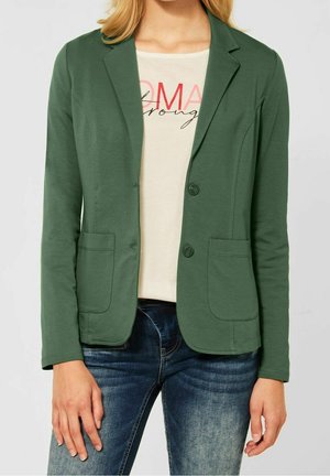 Kvinde iført en mørkegrøn blazer med knapper over en hvid grafisk t-shirt og falmede blå jeans, vist fra nakke til hofter.