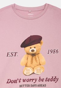 T-shirt rosa claro com uma graphic de um ursinho de pelúcia a usar uma boina escura. O texto inclui "Não te preocupes, sê teddy" e "EST. 1986." Material de algodão macio.