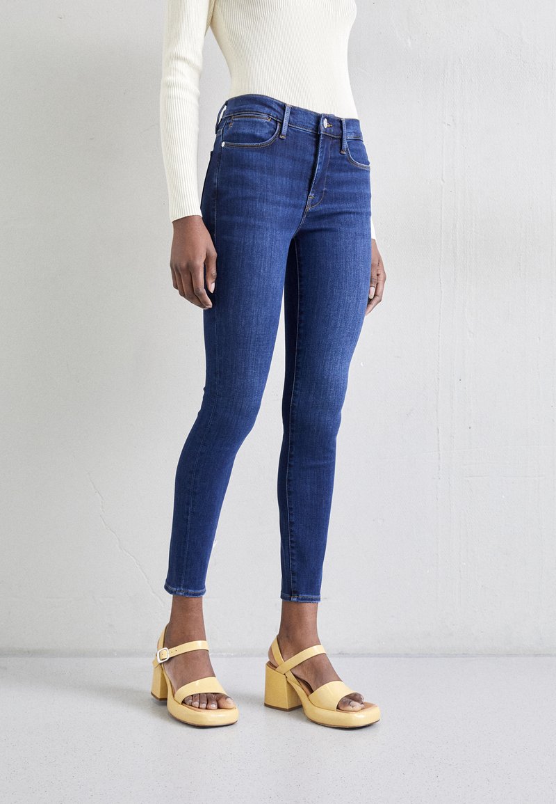 Jeans skinny en denim bleu foncé, avec un style classique à cinq poches, et une silhouette ajustée. Associés à des sandales à plateforme jaunes.