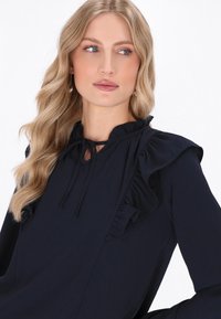 Blusa a maniche lunghe blu navy con dettagli arricciati sulle spalle e sul colletto, caratterizzata da una chiusura a fiocco e una texture di tessuto liscia.