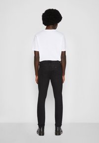 Personne aux cheveux courts et bouclés portant un t-shirt blanc, un jean noir ajusté et des bottes noires, debout devant un fond gris uni.