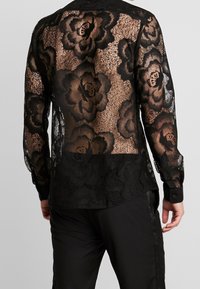 Camicia in pizzo nero con motivo floreale, maniche lunghe e polsini con bottoni neri. Design semi-trasparente con orlo inferiore nero solido.