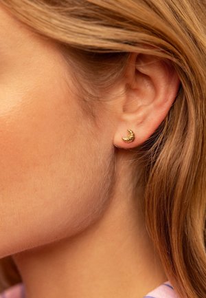 Primer plano de la oreja izquierda con un pendiente corto en forma de media luna dorada, cabello castaño claro recogido detrás de la oreja y piel lisa visible.