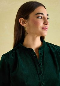 Grün Samtshirt mit Stehkragen, Vorderknöpfen und dezenten Falten an den Schultern. Model trägt goldene Creolen.