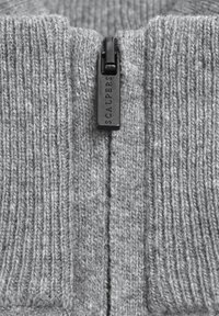 Grau gestrickter Pulloverstoff mit vertikalen Rippungen, ausgestattet mit einem schwarzen Reißverschluss und einem an dem Reißverschluss befestigten Etikett mit der Bezeichnung "SCALPERS".