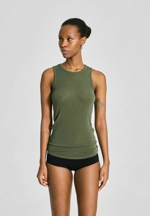 Γυναίκα με αμάνικο τοπ λαδί και μαύρο σορτς, στέκεται και κοιτάζει αριστερά, σε απλό φωτεινό φόντο.