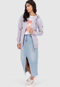 Felpa zip-up lavanda con cappuccio e coulisse, indossata sopra una maglietta bianca con grafica, abbinata a una gonna di jeans chiara con spacco frontale. Sneakers bianche.