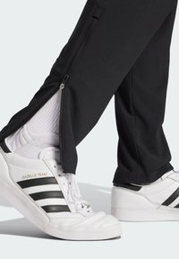 Pantalones deportivos negros con cremalleras laterales combinados con zapatillas blancas que presentan franjas negras y el texto dorado "GAZELLE TEAM". Suelas de goma texturizada.