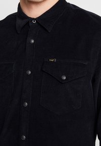 Czarna koszula z corduroy z guzikami na przodzie, kołnierzykiem i dwoma kieszeniami na piersi. Wyposażona w metalowe guziki oraz małą metkę z logo.