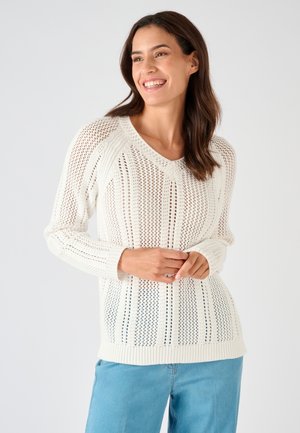 Femme souriante aux cheveux bruns mi-longs portant un pull en maille blanc et un jean bleu clair sur un fond uni.
