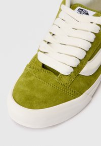 Sneaker verde in suede con lacci bianchi e accenti laterali, punta arrotondata, suola in gomma e dettagli di cucitura visibili.