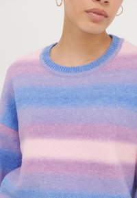 Femme portant un pull à rayures pastel avec des bandes horizontales roses, violettes et bleues et un col côtelé bleu, avec une boucle d'oreille en or visible.