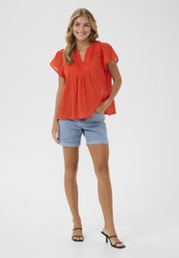 Haut rouge à manches courtes avec décolleté en V et détail froncé, associé à un short en denim bleu clair retroussé à l'ourlet et des sandales à talons hauts noires.