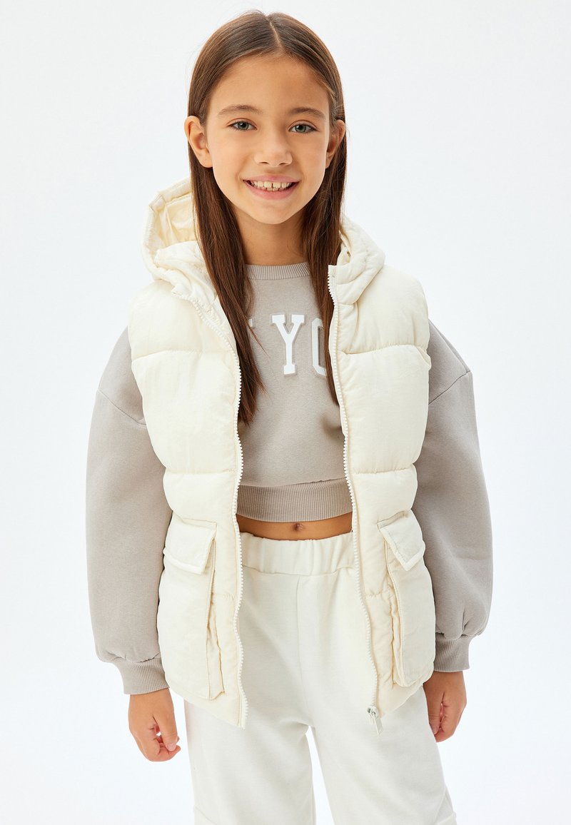 Puffväst i off-white med huva, framdragkedja och två sidofickor. Bärs över en ljusgrå kortärmad sweatshirt och ljusa byxor.