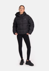 Giacca a vento nera con cappuccio, caratterizzata da un design trapuntato e un piccolo logo bianco sul lato sinistro del petto. Indossata con leggings neri e sneakers.