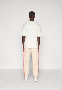 Hellbeiges Oversize-T-Shirt mit roten Schulterakzenten, kombiniert mit hellbeigen Jogginghosen mit roten Seitenstreifen. Glatte Stofftextur.