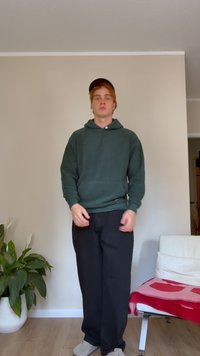 Hanorac verde închis cu glugă, croială lejeră, pantaloni negri, papuci gri, o persoană stând în picioare lângă un perete neutru, cu o plantă în apropiere.