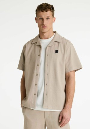 Mladý muž so krátkymi kučeravými vlasmi, oblečený v béžovej krátkorukávovej košeli na gombíky cez biele tričko, na jednoduchom pozadí.