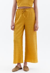 Pantalons larges jaunes, texturés, avec une taille élastique et un cordon de serrage. Associés à des chaussures à enfiler en tissu tissé.