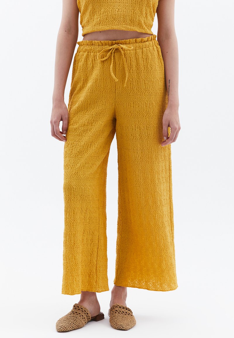 Pantalons larges jaunes, texturés, avec une taille élastique et un cordon de serrage. Associés à des chaussures à enfiler en tissu tissé.