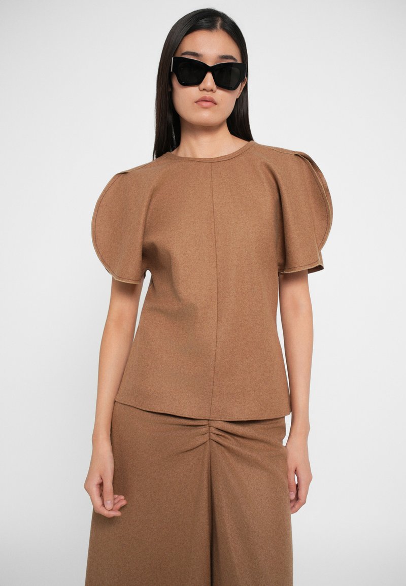 Top marrone con texture e maniche a sbuffo, abbinato a una gonna arricciata coordinata. La modella indossa occhiali da sole neri con forma angolare.