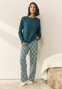 Langärmliges tealfarbenes Oberteil, kombiniert mit grünen gestreiften Schlafanzughosen mit geometrischen Mustern. Das Modell steht auf einem hellen Teppich.