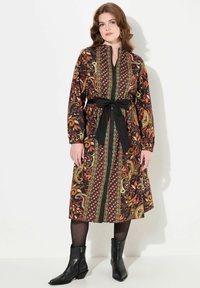 Lungo abito button-up nero con un motivo paisley multicolore, dotato di una cintura nera, maniche lunghe e una vestibilità rilassata. Stivaletti neri alla caviglia.