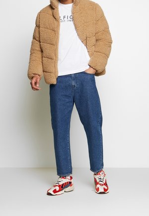 Chaqueta beige de felpa con cierre, camiseta blanca con texto, jeans de pierna recta azules y zapatillas con patrón rojo y blanco.