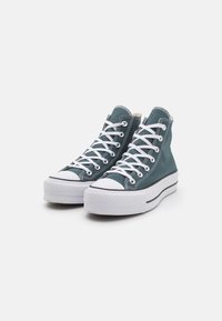 Kotníkové tenisky z tyrkysového plátna s bílými tkaničkami, gumovou špičkou a tlustou bílou podrážkou s černým pruhem. Klasický design s prošitím jako detaily.