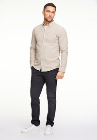 Camicia button-up beige chiaro con tasca frontale, abbinata a pantaloni blu scuro e sneakers bianche. Tessuto liscio, vestibilità su misura.