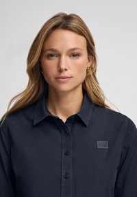 Camicia button-up di un blu navy scuro realizzata in tessuto liscio, con colletto strutturato, bottoni frontali e una piccola patch con logo sul petto.