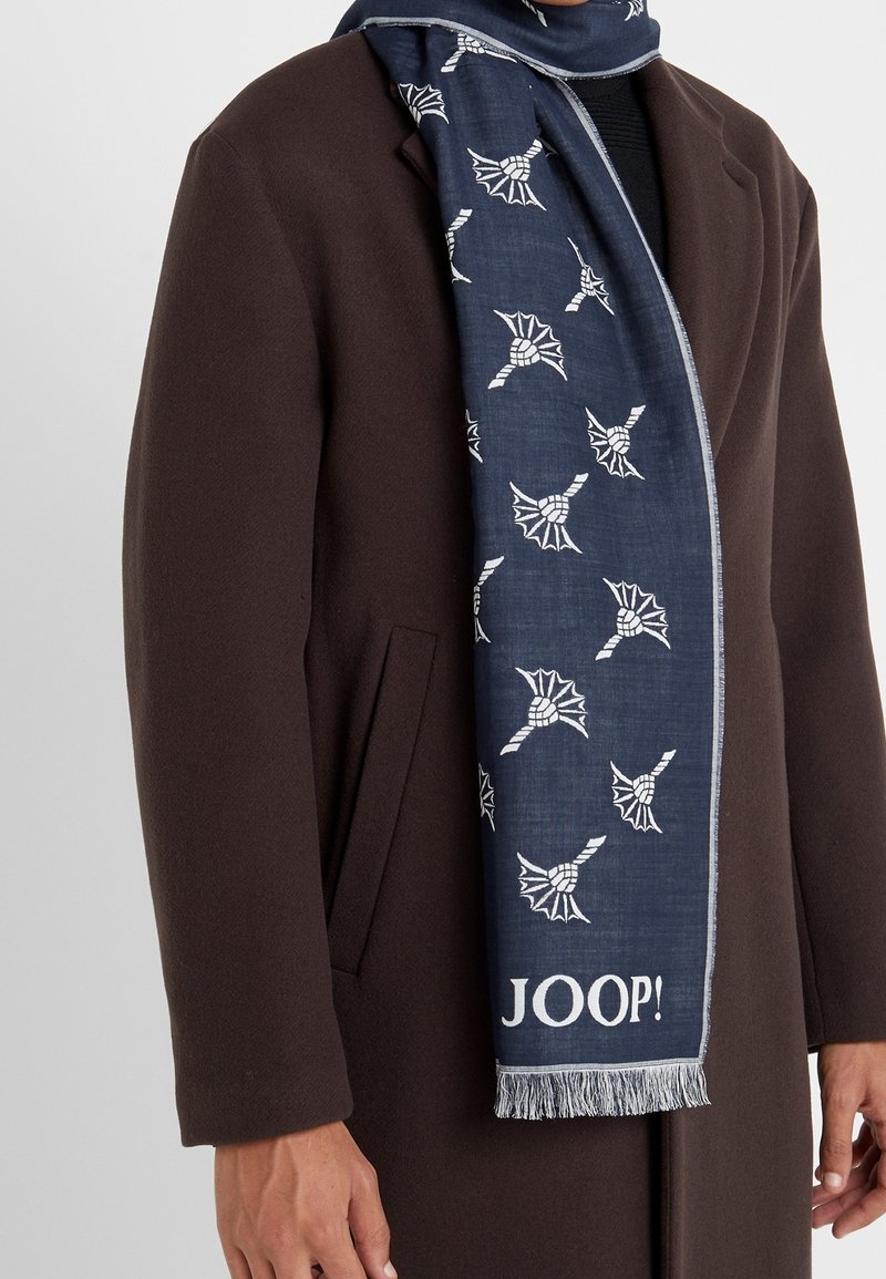 Homme portant un manteau marron foncé et une écharpe bleu marine avec des motifs de feuilles blanches et le texte "JOOP!" près du bord à franges.