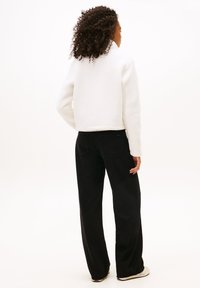White fleece jacket with a high collar, paired with loose-fitting black pants and light-colored shoes. View from the back.