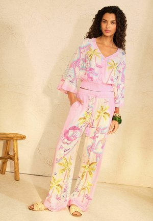 Vrouw draagt een pastelroze bijpassende top en wijde broek met tropische palmbomen en flamingoprint, staat naast een houten kruk.