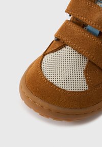 Καφέ παπούτσι από σουέτ με τμήμα από ύφασμα διχτυού και δύο ιμάντες με βέλκρο. Η σόλα είναι ελαφρώς διαφανής και υφή για πρόσφυση.