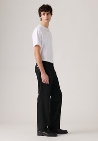 Man wearing white short-sleeve shirt, black pants, and black shoes standing sideways against plain background.