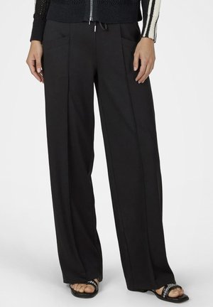 Schwarze Hosen mit weitem Bein, vorn mit Taschen und sichtbaren Nähten, kombiniert mit einem schwarz-weiß gestreiften Pullover mit Ärmeln und schwarzen Sandalen mit Verzierungen.