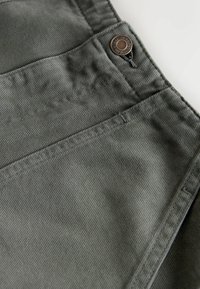 Zbliżenie na szare spodnie dżinsowe ukazujące przednią kieszeń, szlufkę na pasek i metalowy guzik z grawerem marki.