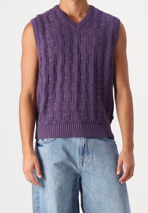 Homme portant un gilet en maille texturée violet sans manches avec col en V, associé à un jean en denim clair, debout devant un fond uni.