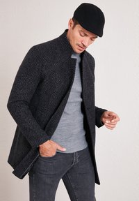 Cappotto nero strutturato con colletto alto, indossato sopra un maglione grigio a coste. Jeans grigi e un berretto nero completano il look.