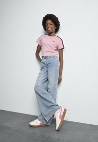 T-shirt courte rose clair avec des accents bordeaux, associée à un jean large bleu clair et des baskets blanches avec des détails roses et bruns.