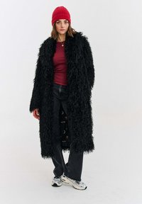 Černý dlouhý kabát z umělé kožešiny, vínově rudý přiléhavý top, tmavé džíny a stříbrné tenisky; na hlavě červená žebrovaná čepice; materiály s texturou a vrstvený vzhled.